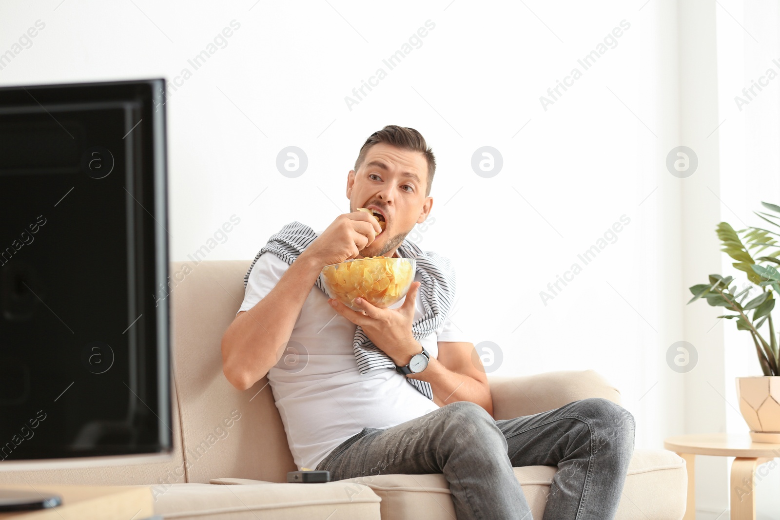 Photo of Man with bowl of potato chips watching TV on sofa in living room