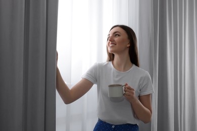Photo of Woman with cup of hot drink opening stylish curtains at home
