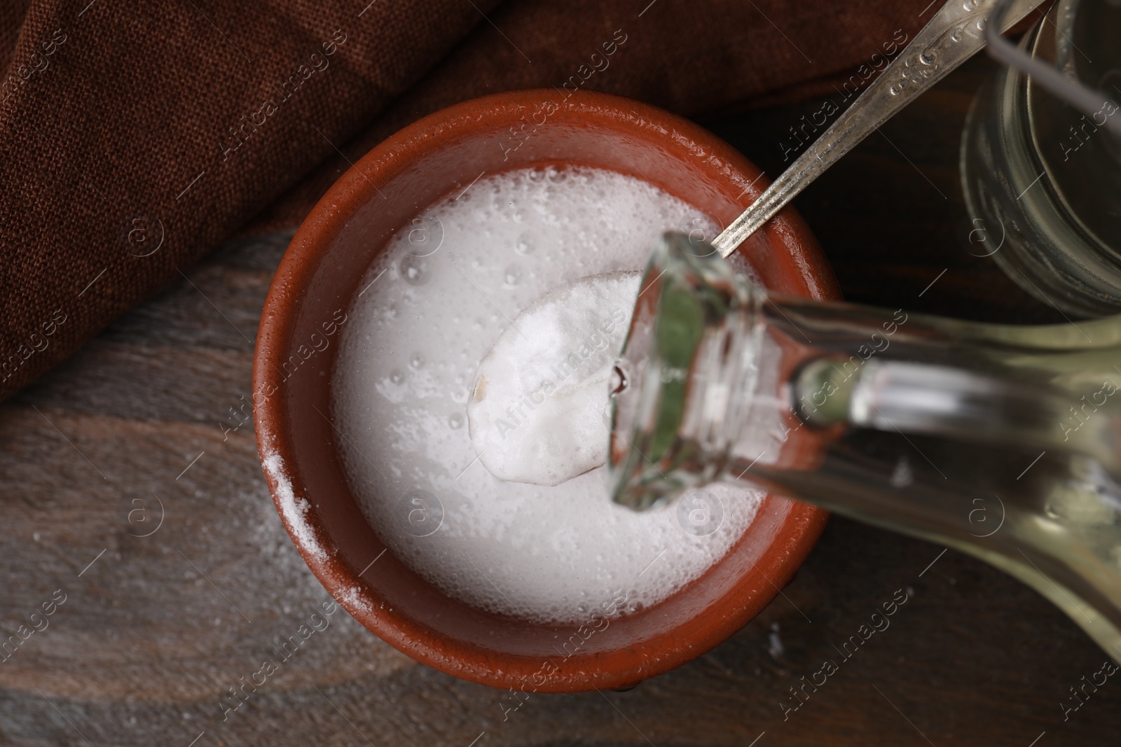 Photo of Pouring vinegar into spoon with baking soda over bowl at wooden table, top view