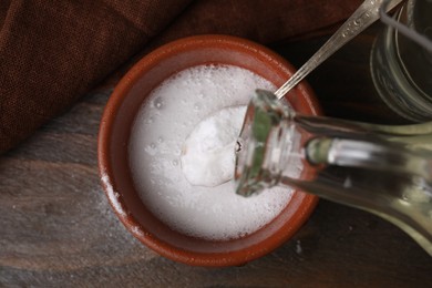 Pouring vinegar into spoon with baking soda over bowl at wooden table, top view