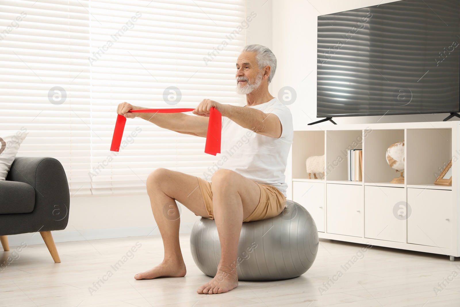 Photo of Senior man doing exercise with elastic resistance band on fitness ball at home