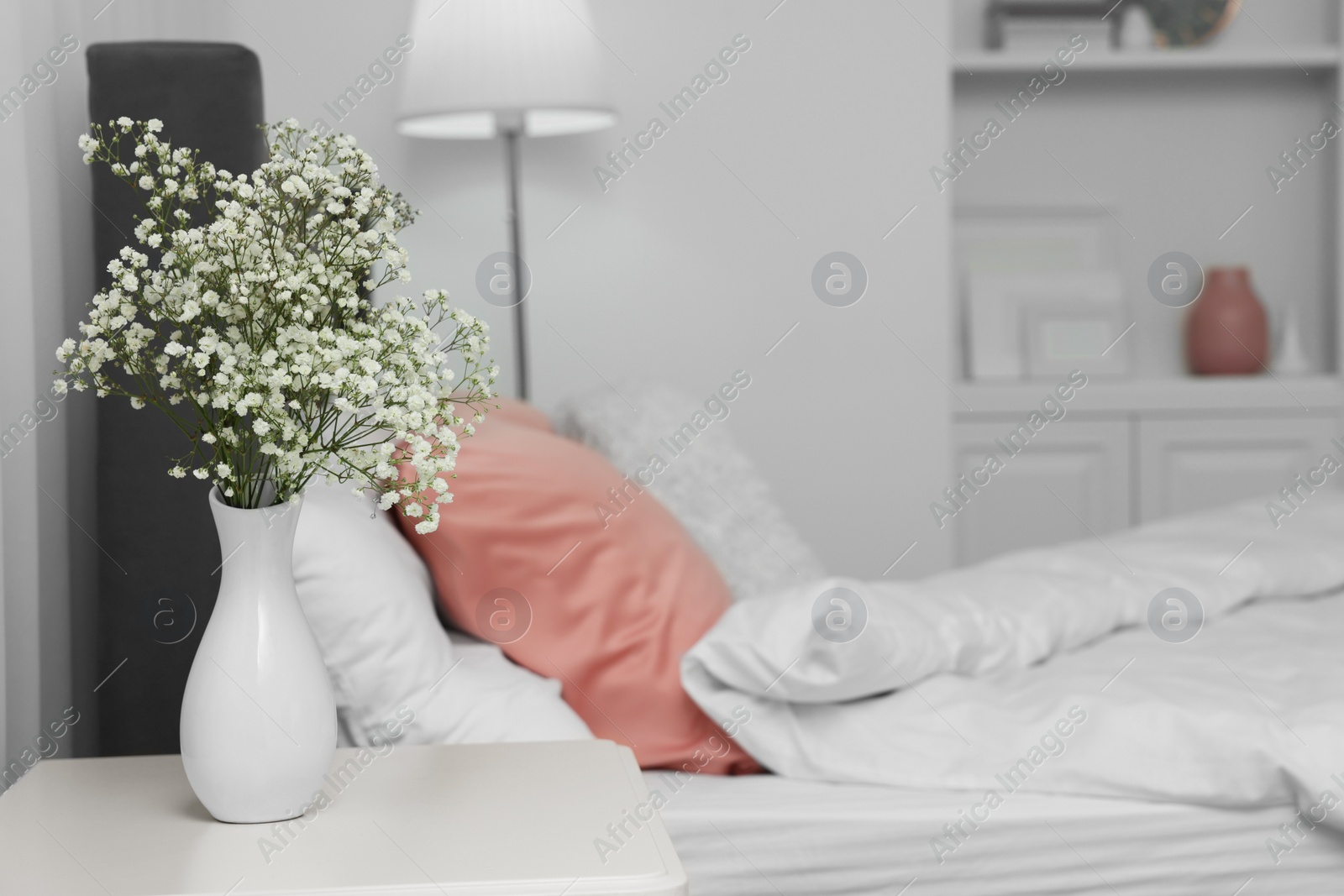 Photo of Vase with beautiful flowers on white bedside table near comfortable bed in room, space for text. Stylish interior