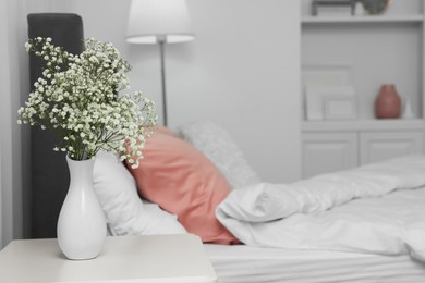 Vase with beautiful flowers on white bedside table near comfortable bed in room, space for text. Stylish interior