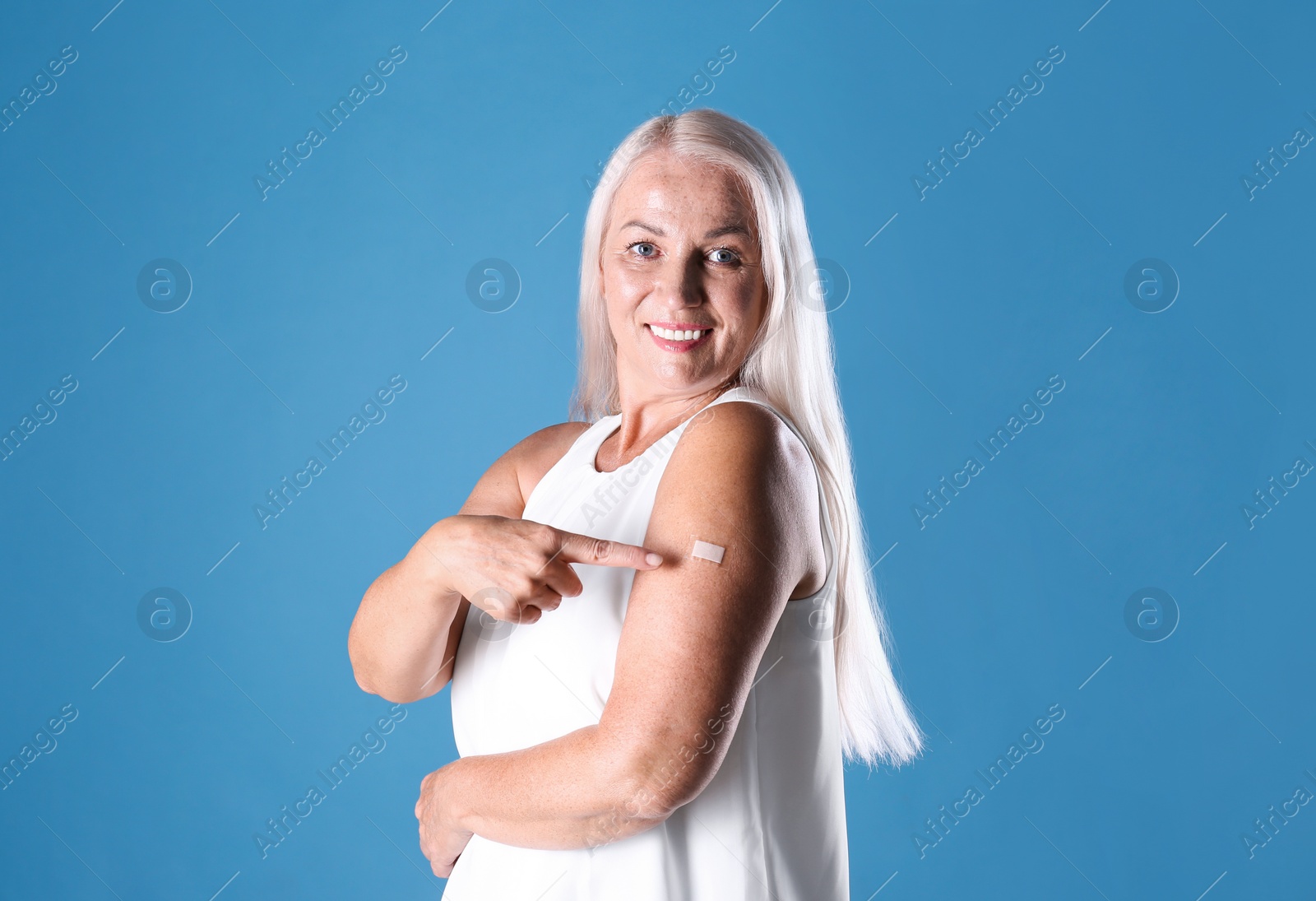 Photo of Happy mature woman pointing at arm with bandage after vaccination on light blue background