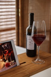 Wooden tray with tablet, glass of wine and bottle on bathtub in bathroom