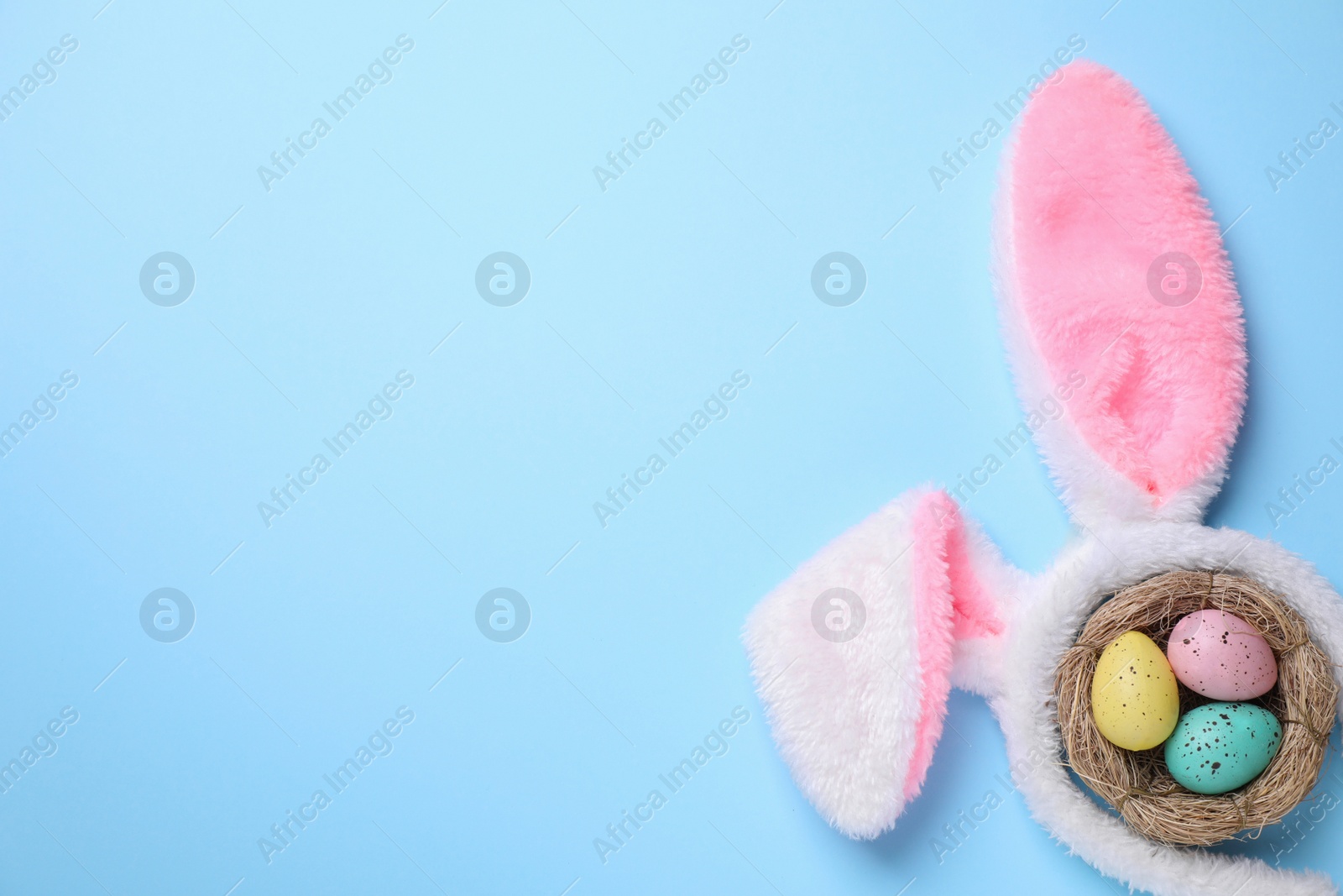 Photo of Headband with bunny ears, painted eggs and space for text on light blue background, flat lay. Easter holiday