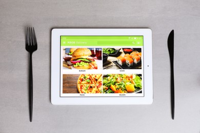 Photo of Modern tablet with open page for online food ordering and cutlery on grey table, flat lay. Concept of delivery service