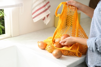 Photo of Woman taking golden onion from mesh tote bag at countertop in kitchen, closeup