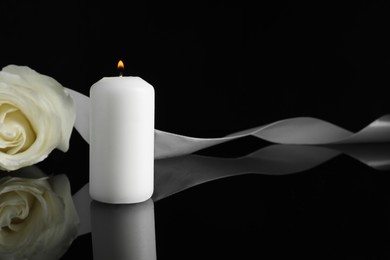 Burning candle, white rose and ribbon on black mirror surface in darkness, space for text. Funeral symbols