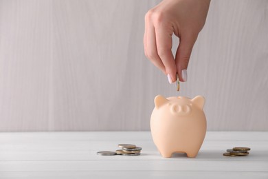 Woman putting coin into piggy bank at white wooden table, closeup with space for text