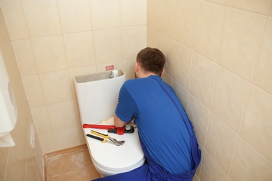 Professional plumber in uniform fixing toilet tank indoors