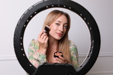 Photo of Beautiful young woman applying blusher with brush near white wall, view through ring lamp