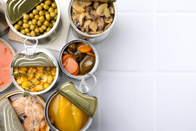 Photo of Open tin cans with different food on white tiled table, flat lay. Space for text