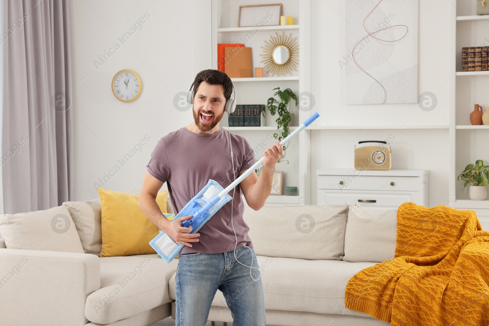 Photo of Spring cleaning. Man in headphones with mop singing while tidying up living room