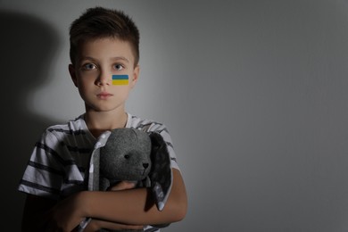 Image of Sad little boy with toy and picture of Ukrainian flag on cheek near light wall, space for text. Stop war in Ukraine