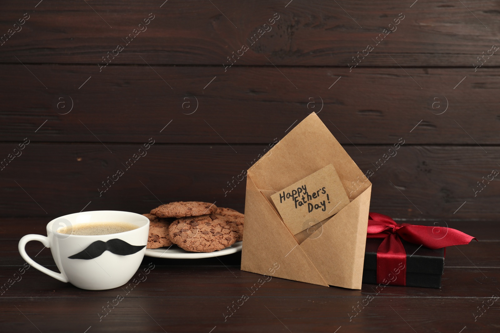 Photo of Card with phrase Happy Father's Day in envelope, cookies, cup with moustache and gift box on wooden table