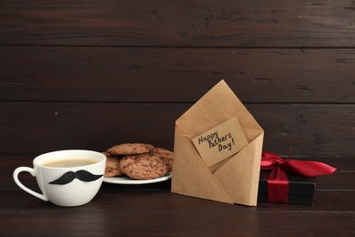 Photo of Card with phrase Happy Father's Day in envelope, cookies, cup with moustache and gift box on wooden table