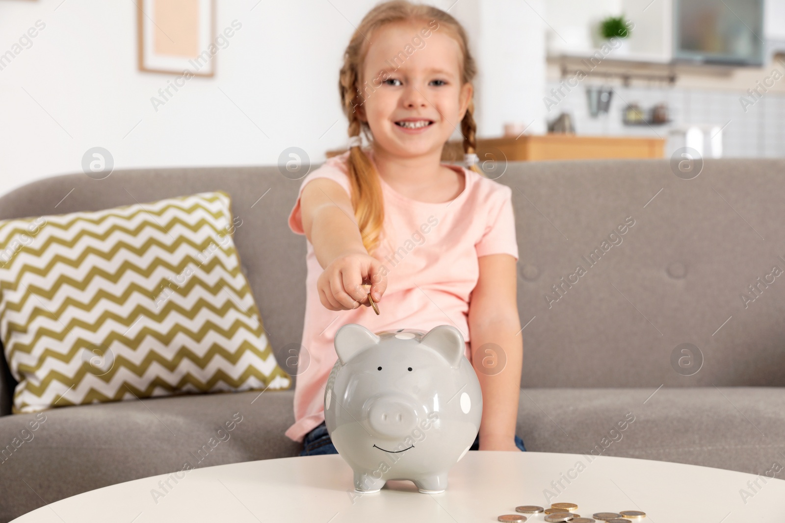 Photo of Cute girl putting coin into piggy bank at table in living room. Saving money