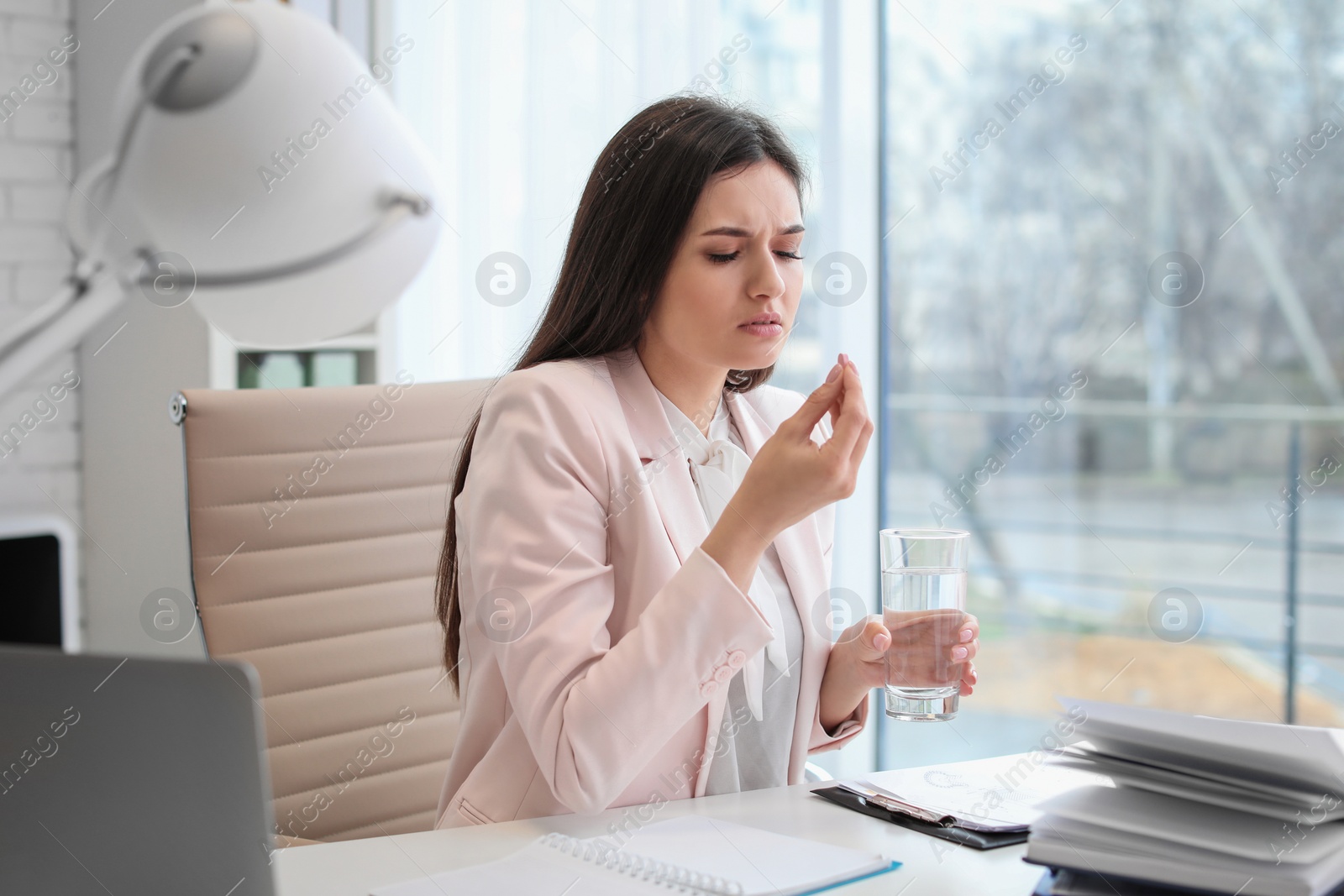 Photo of Young woman taking pill against headaches while sitting at table in office