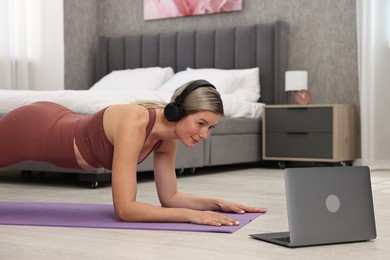 Photo of Online fitness trainer. Woman in headphones doing plank exercise near laptop at home