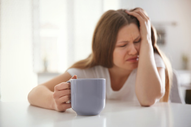 Sleepy young woman with cup of drink at home in morning, focus on hand