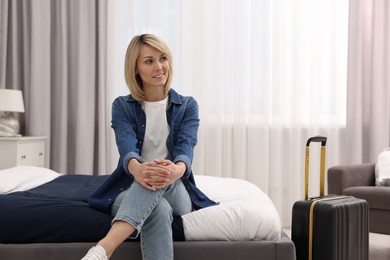 Photo of Smiling guest with suitcase relaxing on bed in stylish hotel room