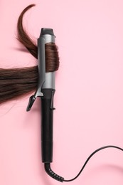 Curling iron with brown hair lock on pink background, top view