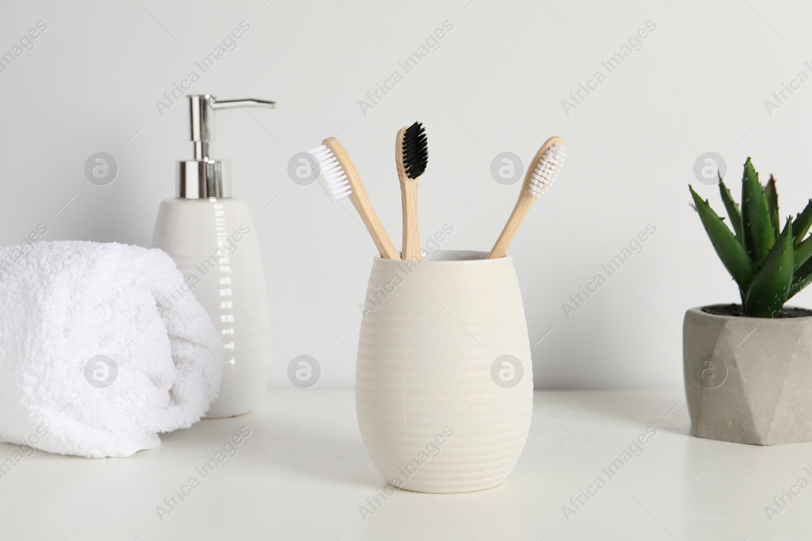 Photo of Bamboo toothbrushes in holder, potted plant, towel and cosmetic product on white countertop