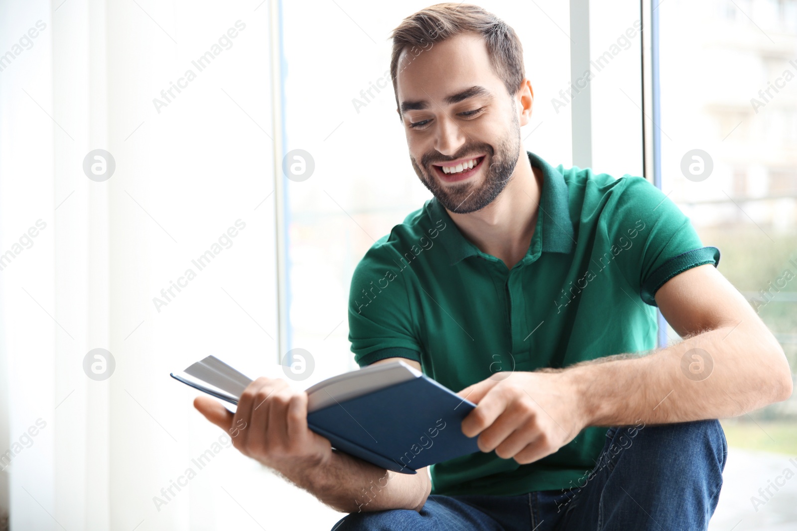 Photo of Young man reading book near window at home