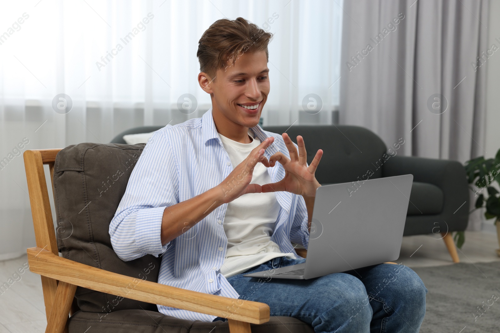 Photo of Happy young man having video chat via laptop and making heart with hands on armchair indoors. Long-distance relationship