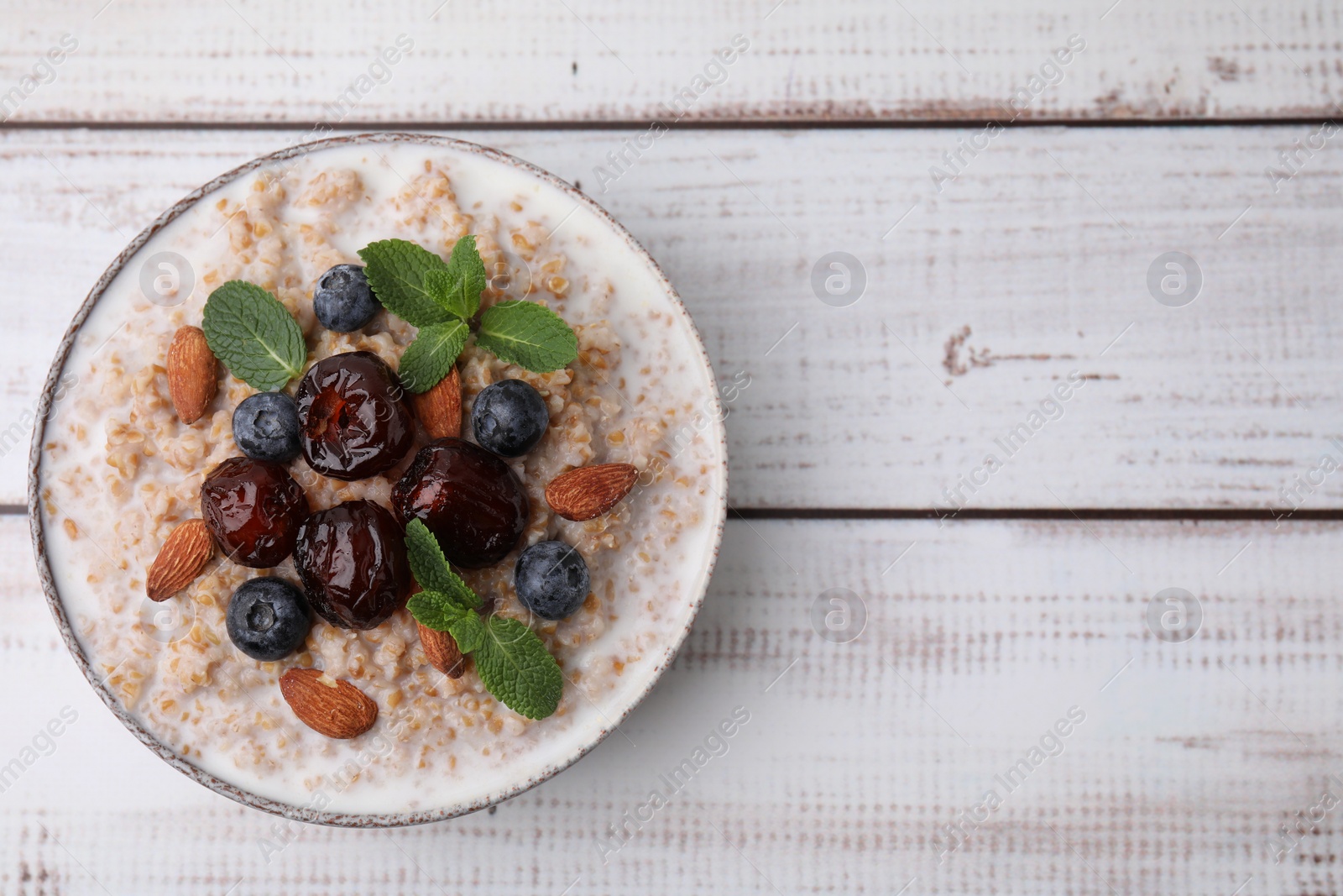 Photo of Tasty wheat porridge with milk, dates, blueberries and almonds in bowl on light wooden table, top view. Space for text