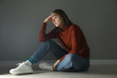 Photo of Sad young woman sitting on floor near grey wall indoors