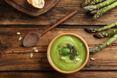 Photo of Delicious asparagus soup in bowl served on wooden table, flat lay