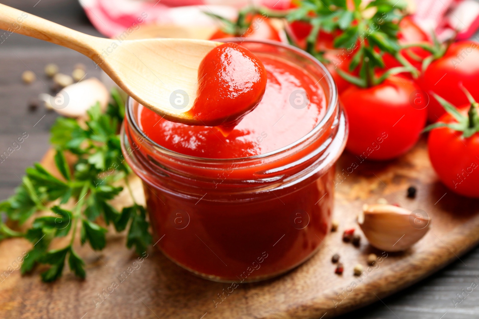 Photo of Jar and spoon with tasty ketchup, fresh tomatoes, parsley and spices on grey wooden table, closeup