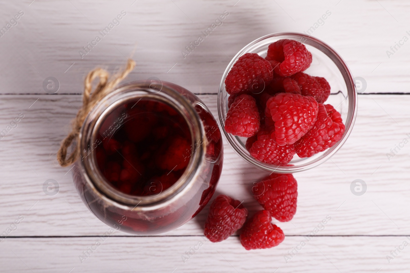 Photo of Jar of tasty canned raspberry jam and fresh berries on white wooden table, flat lay