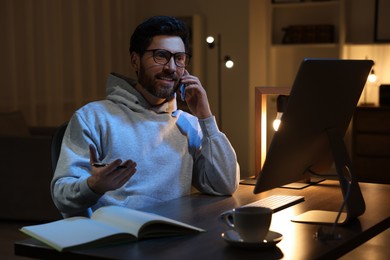 Photo of Home workplace. Man talking on smartphone while working with computer at wooden desk at night