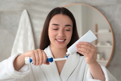 Photo of Young woman squeezing toothpaste from tube onto electric toothbrush in bathroom