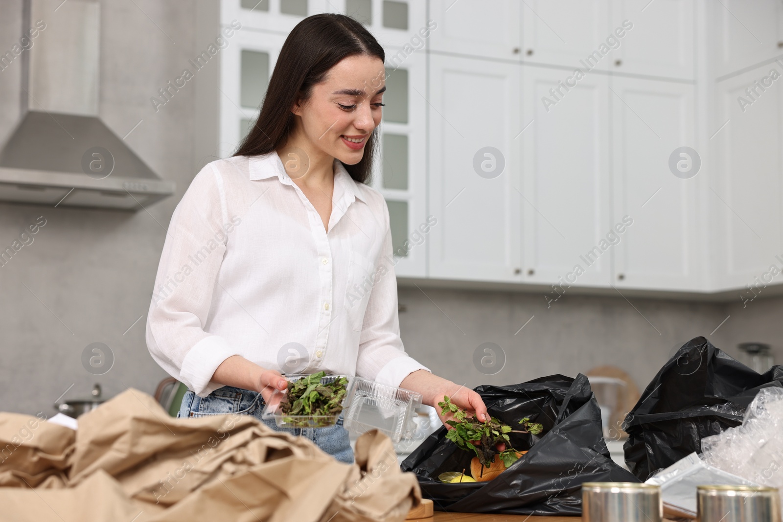 Photo of Garbage sorting. Woman putting food waste into plastic bag at table in kitchen