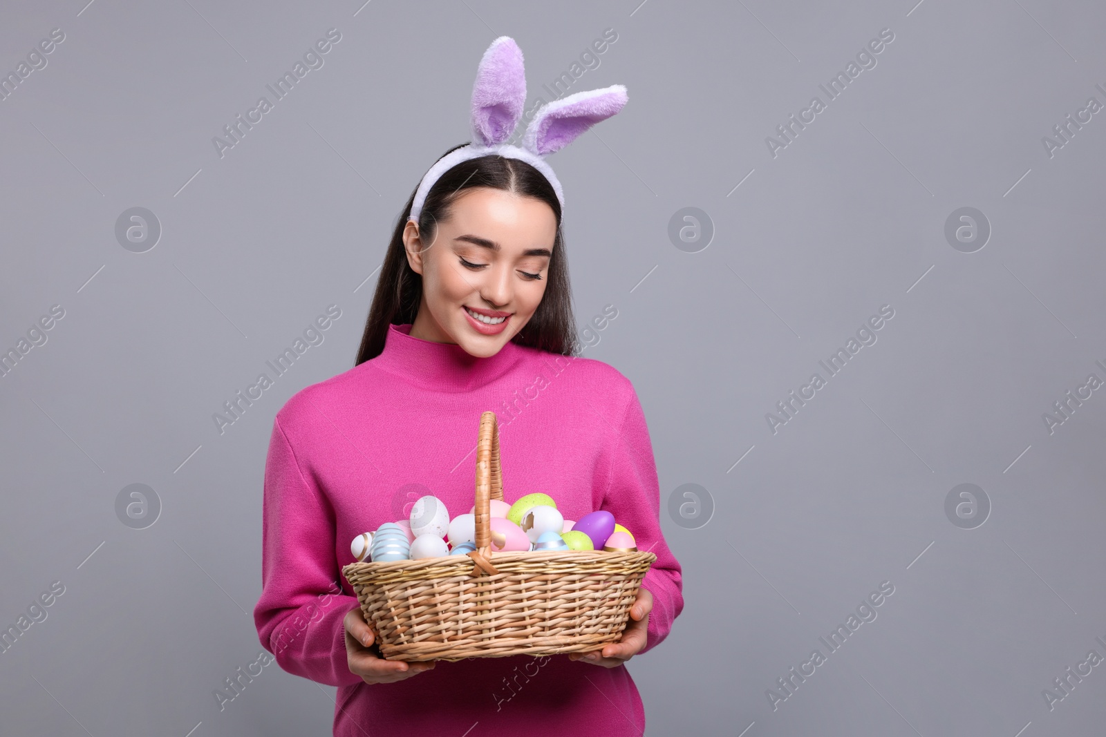 Photo of Happy woman in bunny ears headband holding wicker basket of painted Easter eggs on grey background. Space for text