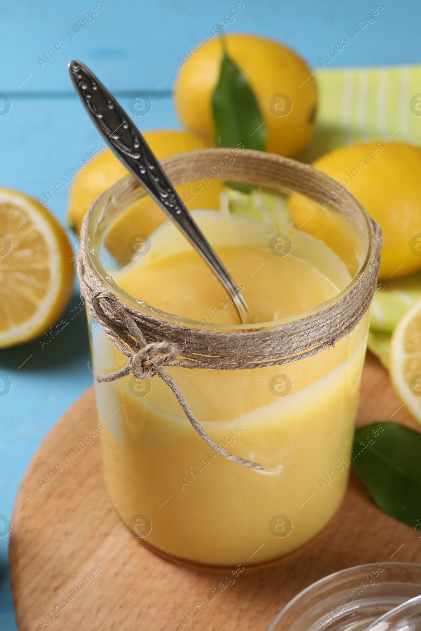 Photo of Delicious lemon curd in glass jar, fresh citrus fruits and spoon on light blue table, closeup