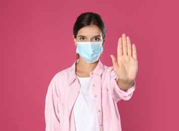 Photo of Woman in protective mask showing stop gesture on pink background. Prevent spreading of coronavirus