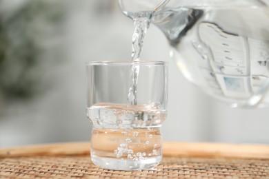 Pouring fresh water from jug into glass at wicker surface against blurred background, closeup