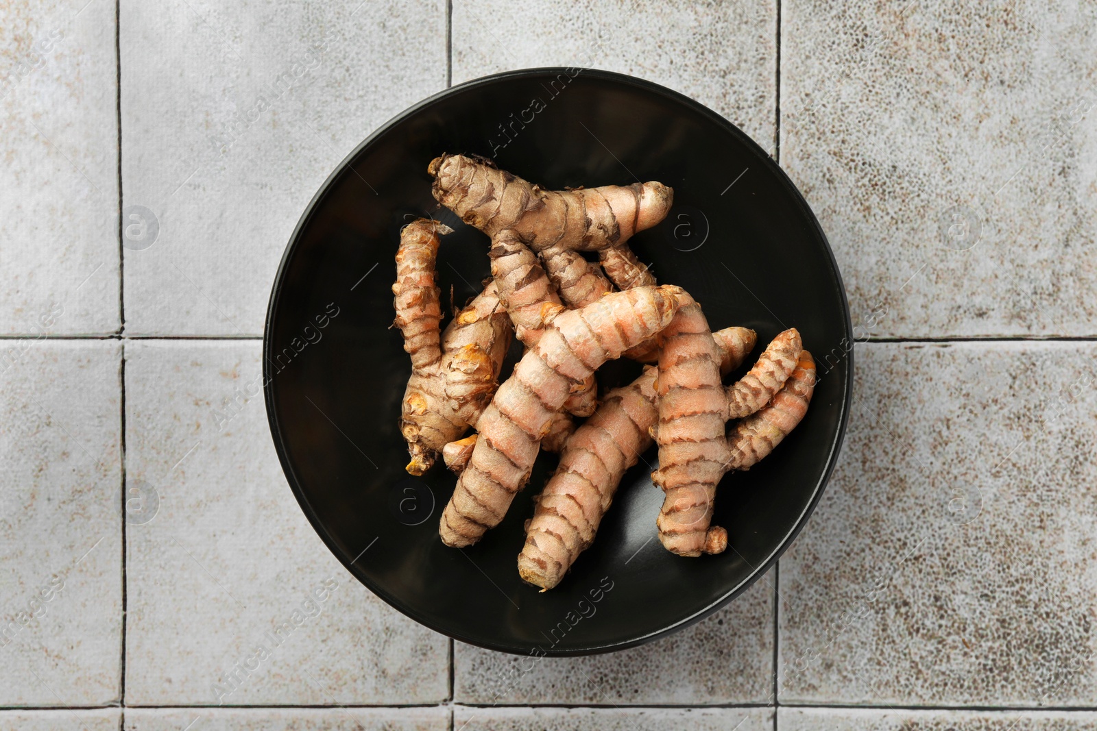 Photo of Bowl with raw turmeric roots on white tiled table, top view