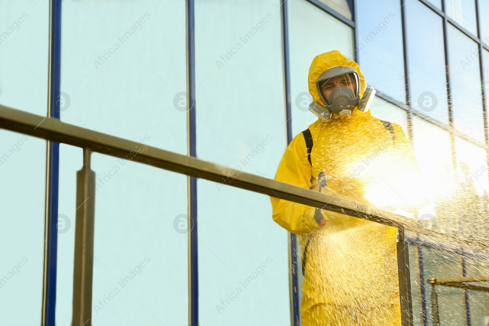 Photo of Person in hazmat suit with disinfectant sprayer cleaning metal railing on city street. Surface treatment during coronavirus pandemic