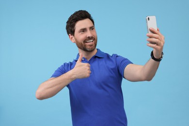 Photo of Smiling man taking selfie with smartphone and showing thumbs up on light blue background