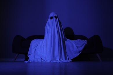 Creepy ghost. Woman covered with sheet sitting on sofa in blue light