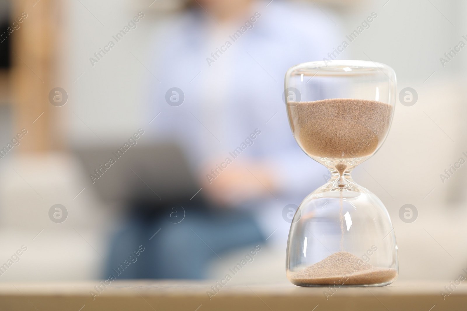 Photo of Hourglass with flowing sand on desk. Woman using laptop indoors, selective focus