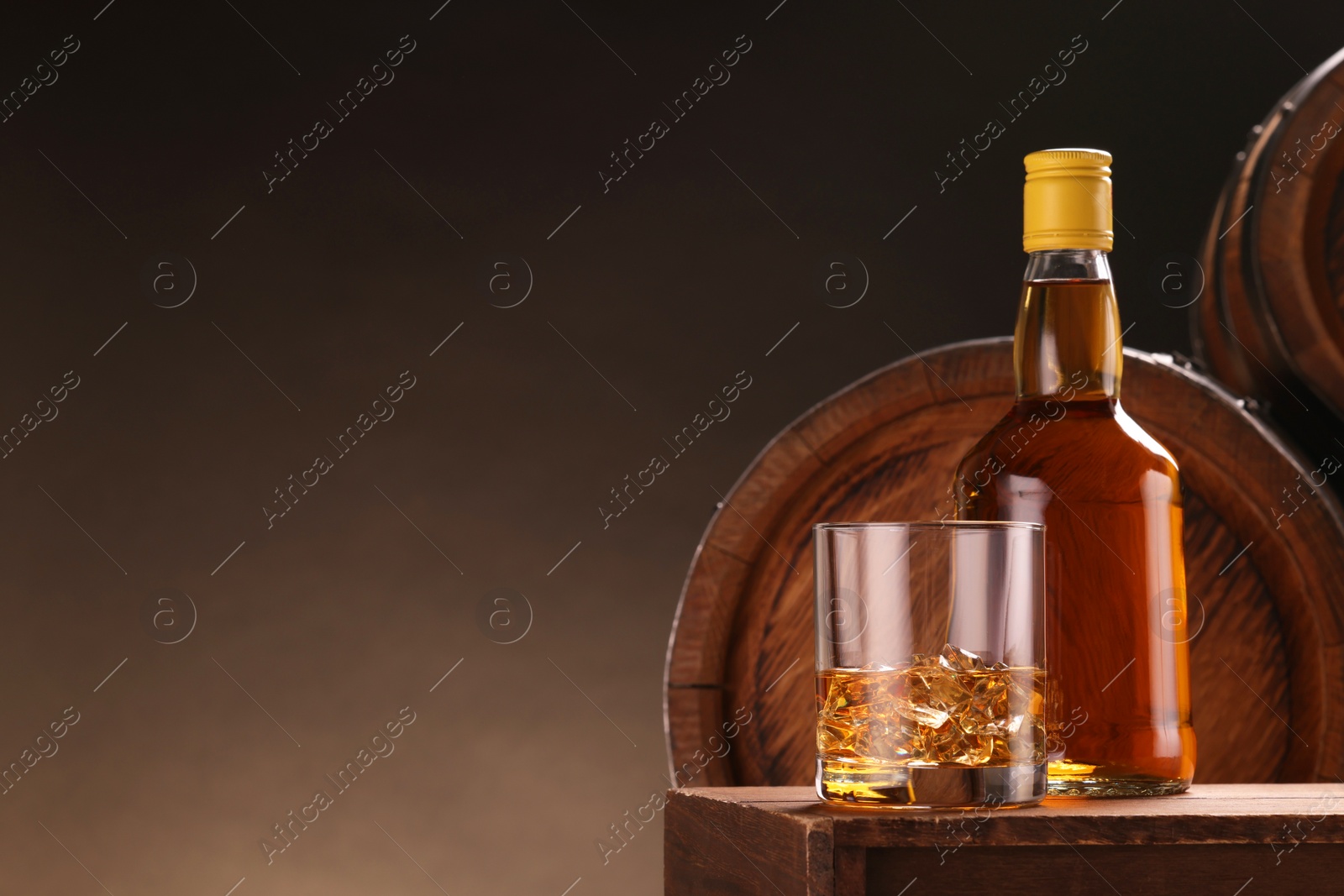 Photo of Whiskey with ice cubes in glass and bottle on wooden table near barrels against dark background, space for text