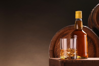 Whiskey with ice cubes in glass and bottle on wooden table near barrels against dark background, space for text