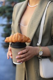 Woman holding tasty croissant and cup of coffee outdoors, closeup